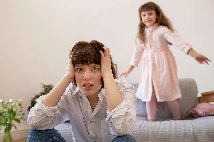 Woman with concerned expression holding her head while her sister plays in a gentle parenting house setting.
