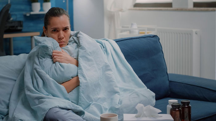 Sick woman wrapped in blanket on couch, with medicine bottles and tissues nearby, showing signs of illness and discomfort.