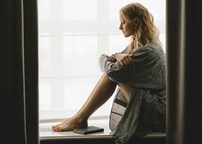 Woman sitting pensively by window, contemplating skipping Thanksgiving after mom’s unhinged text causes embarrassment. Woman sitting pensively by window, contemplating skipping Thanksgiving after mom’s unhinged text causes embarrassment.