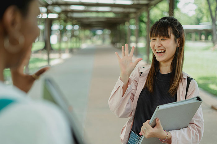 Young woman holding a folder and smiling, waving at a friend in an outdoor walkway, illustrating the impact of lies on lives.