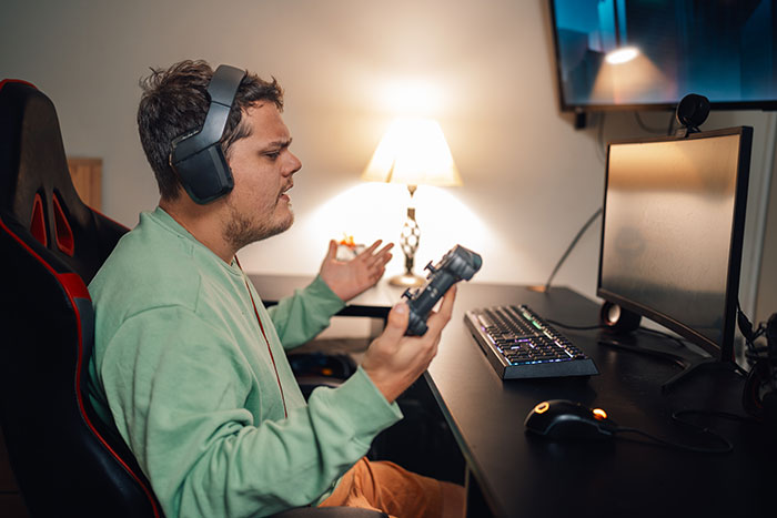 Teen gamer frustrated sitting on broken chair, wearing headset and holding controller in front of computer setup at home.