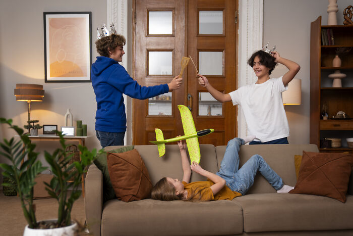 Three children playing with toy swords and a glider airplane on a couch during a family visit at home.
