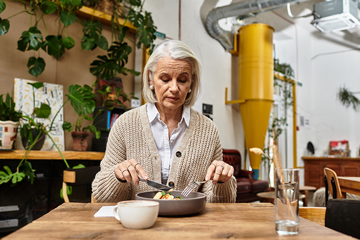 Older woman sitting at a table with a plate of food, appearing thoughtful in a cozy, plant-filled dining space. Older woman sitting at a table with a plate of food, appearing thoughtful in a cozy, plant-filled dining space.