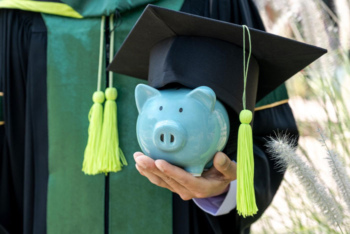 Person in graduation gown holding piggy bank with graduation cap, symbolizing college fund and savings.