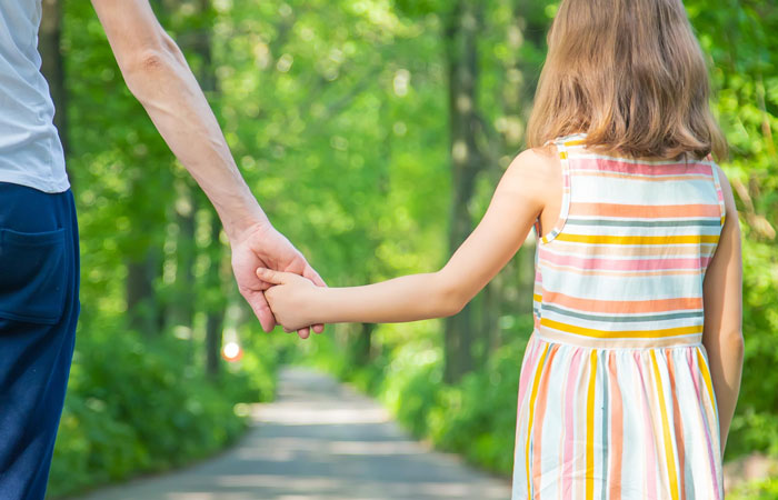 Child holding hands with adult in a park, representing stepdad trying to connect while bio dad remains in kids&rsquo; hearts.