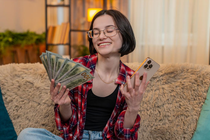 Young woman smiling and holding cash and smartphone indoors, illustrating pregnant SIL paying nieces for help scenario. Young woman smiling and holding cash and smartphone indoors, illustrating pregnant SIL paying nieces for help scenario.