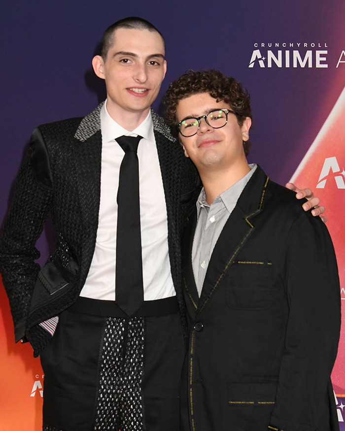 Stranger Things actor Gaten Matarazzo wearing glasses, standing with co-star at a formal event with purple backdrop.