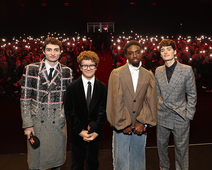 Stranger Things actor Gaten Matarazzo stands with co-stars at a live event with audience lights in the background.