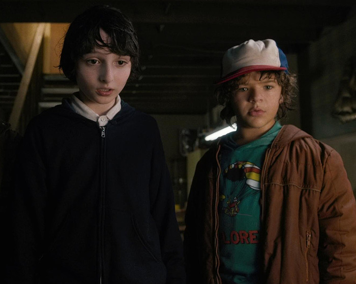 Two Stranger Things actors, including Gaten Matarazzo, standing together in a dimly lit room during a scene.