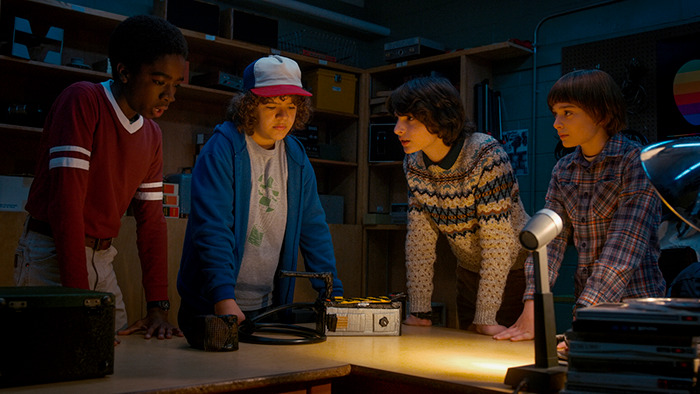 Four Stranger Things actors, including Gaten Matarazzo, gather around a table in a dimly lit room.