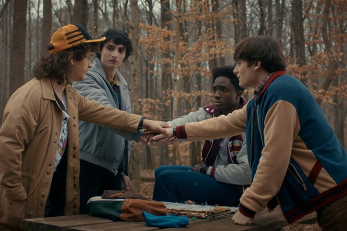 Stranger Things actor Gaten Matarazzo with co-stars outdoors in autumn forest, sharing a team moment around a picnic table.