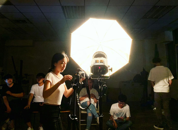 Young woman adjusting camera lighting setup in a dim room while others sit around, illustrating modern movies millennial gray look.