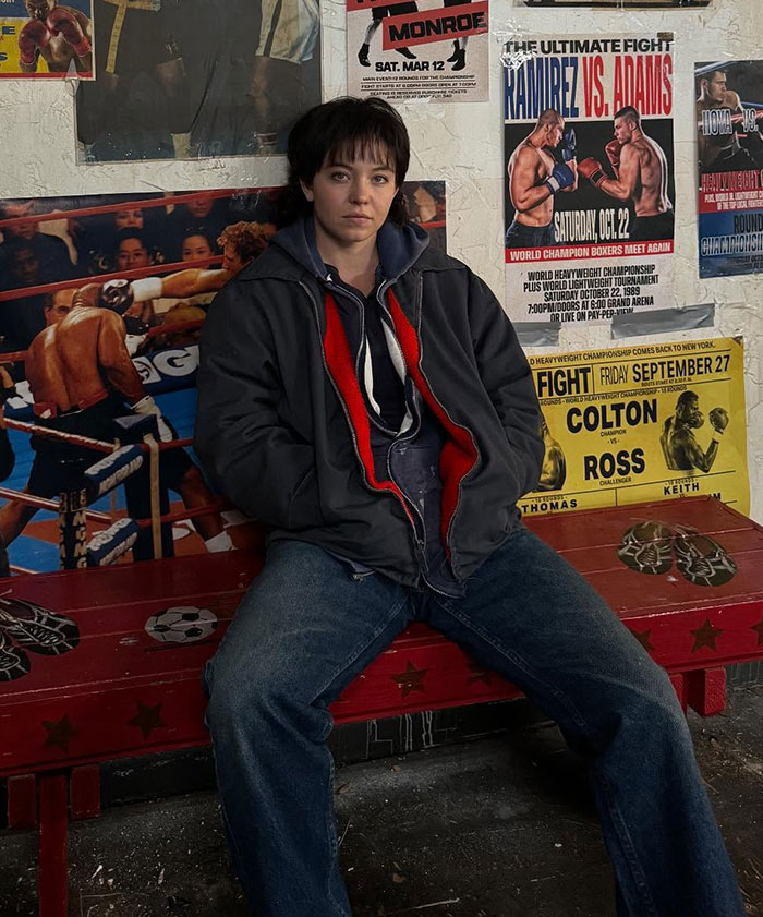 Plus-size model sitting in casual clothes against a wall with boxing posters, relating to Sydney Sweeney body comments. Plus-size model sitting in casual clothes against a wall with boxing posters, relating to Sydney Sweeney body comments.