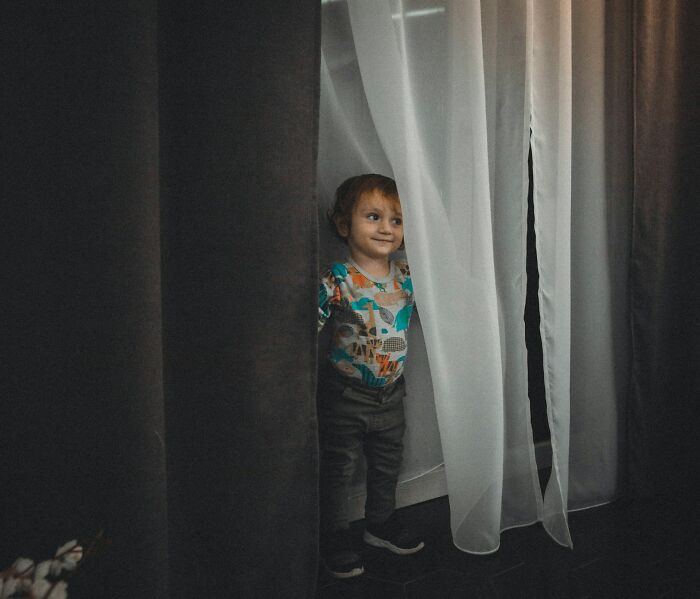 Toddler peeking out from behind curtains with a curious expression, showing kids doing something weird passed down from parents.
