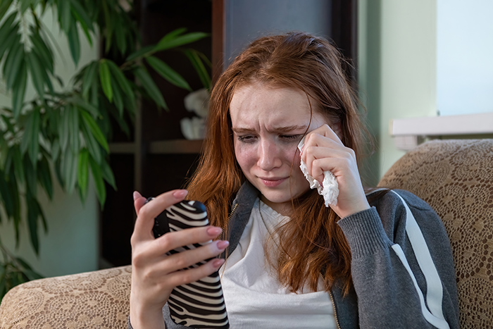 Sad young woman wiping tears while holding phone, reflecting sibling refuse newborn nephew care emotional distress.