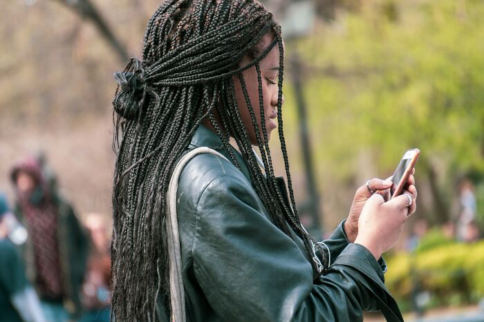 Young woman with long braids wearing a leather jacket, focused on her phone, showing subtle signs of cheating behavior