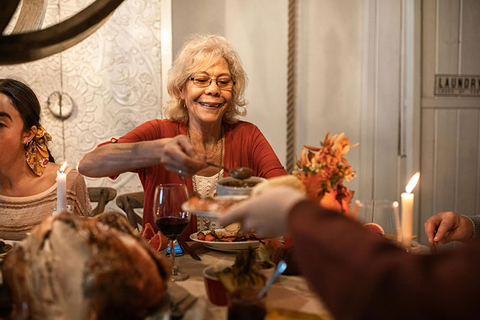 Elderly woman serving food at a Thanksgiving dinner table surrounded by family, highlighting hosting pet peeves.