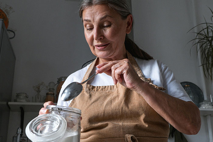 Older woman in kitchen wearing apron, holding a spoon and jar, preparing for Thanksgiving hosting pet peeves.