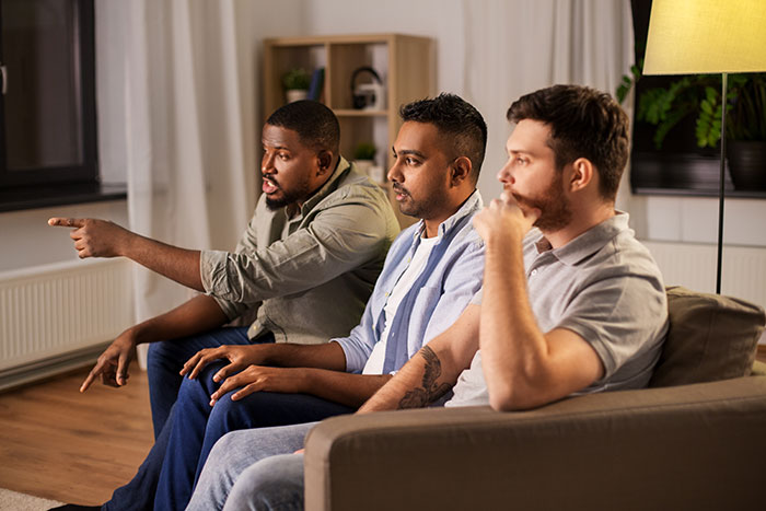 Three men sitting on a couch discussing Thanksgiving hosting pet peeves with focused and engaged expressions.