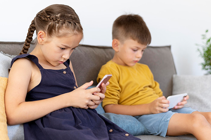 Two children sitting on a couch focused on their smartphones, illustrating concerns about parents posting children online. Two children sitting on a couch focused on their smartphones, illustrating concerns about parents posting children online.