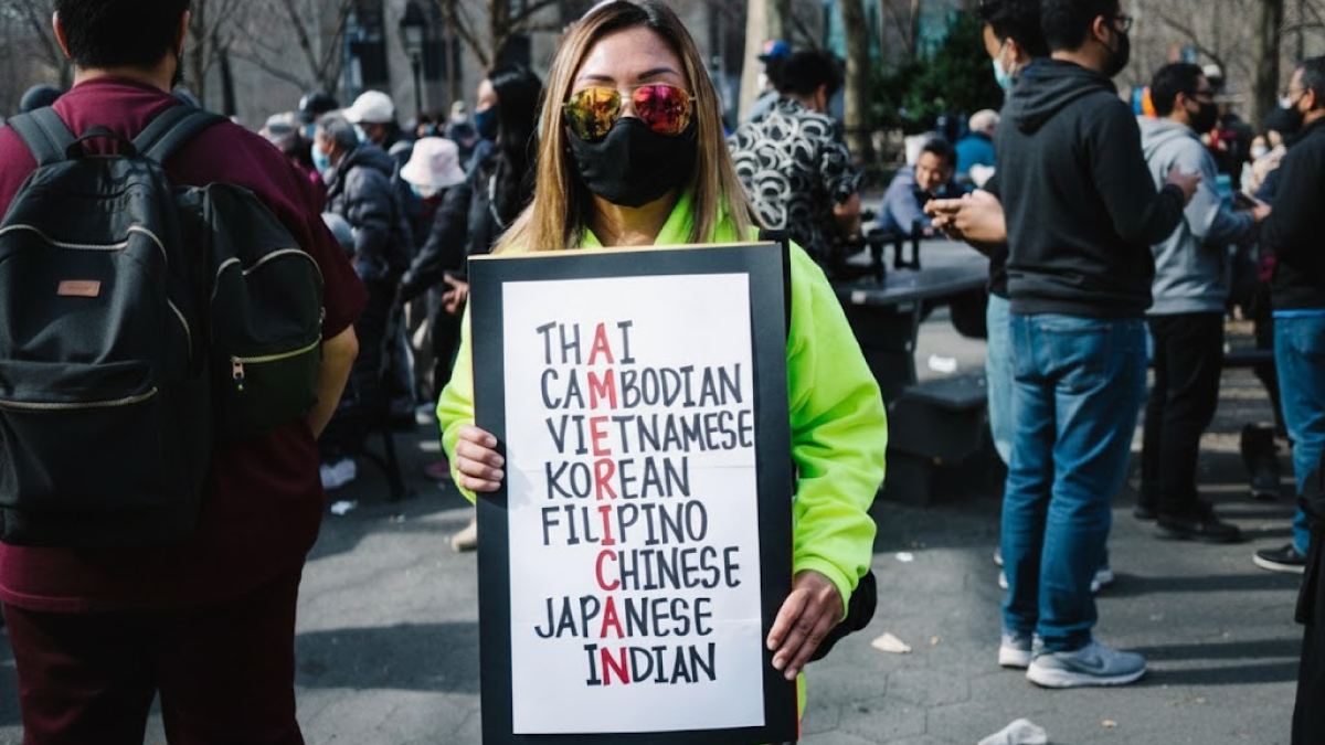 Person wearing sunglasses and a mask holding a sign listing Asian ethnicities at an outdoor event about Asian stereotypes.