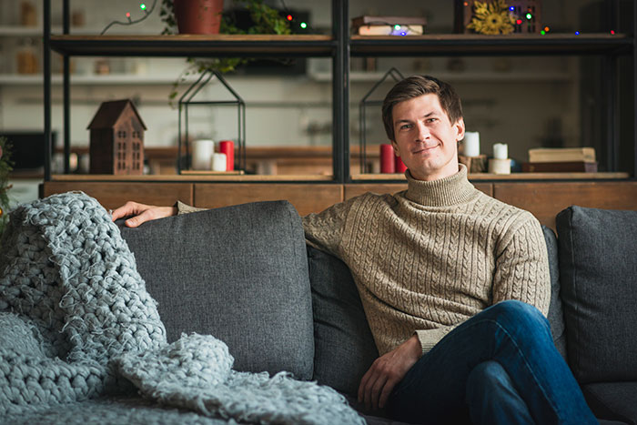 Man in a tan sweater sitting on a gray couch at home, representing men anonymously revealing personal thoughts.