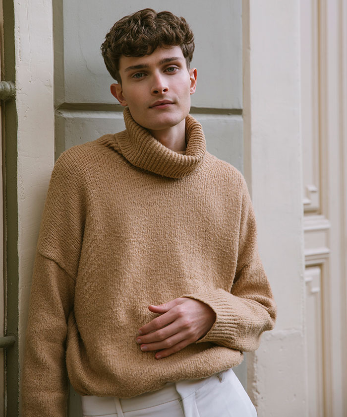 Young man wearing a beige sweater posing thoughtfully against a wall, representing men anonymously revealing secrets.