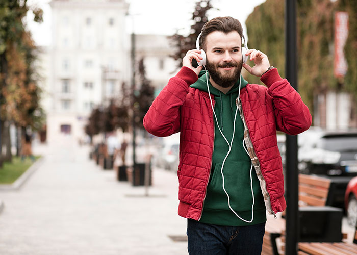 Young man wearing a red jacket and green hoodie, listening to music with headphones on an urban street.