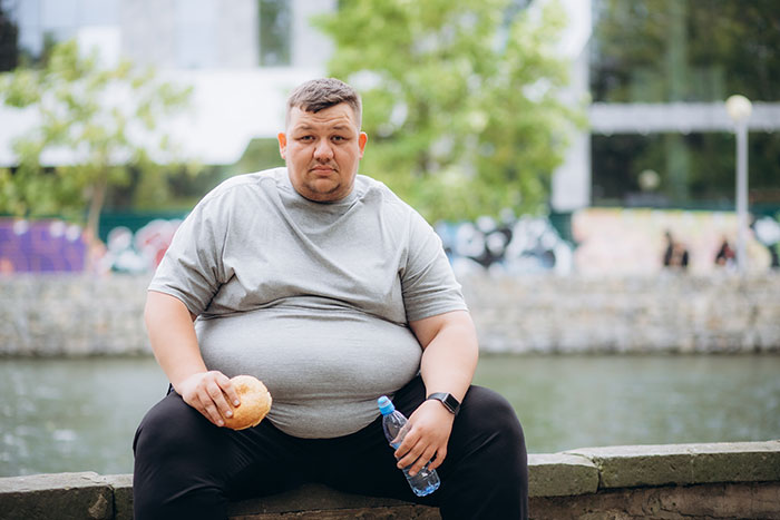 Man sitting outdoors by water holding a sandwich and water bottle, representing metal detectors at school topic.