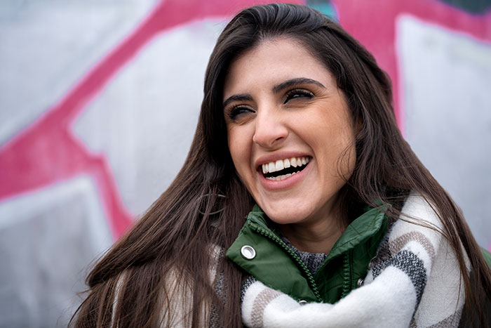 Smiling young woman outdoors wearing a winter coat and scarf with a colorful graffiti wall blurred in the background.