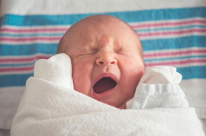 Newborn wrapped in a hospital blanket, yawning while resting peacefully in a bright medical setting.