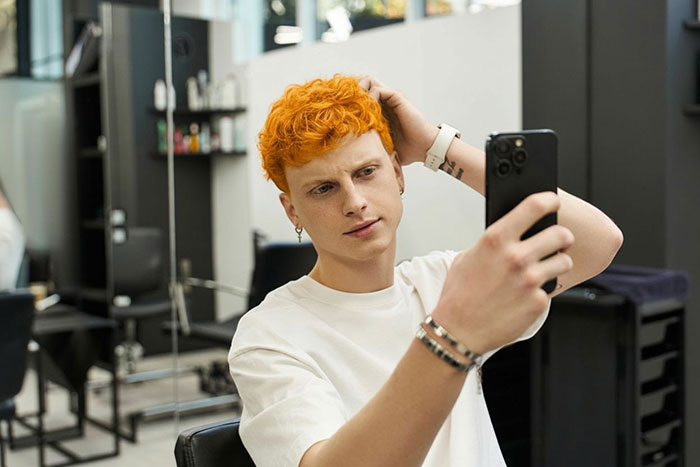 Young man with freshly dyed orange hair taking a selfie in a salon chair, capturing the classmate drama look.