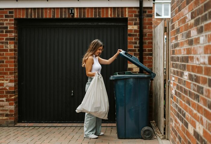 Woman holding a trash bag lifting the lid of a blue garbage bin outside a brick house showing garbage men finds on the job.