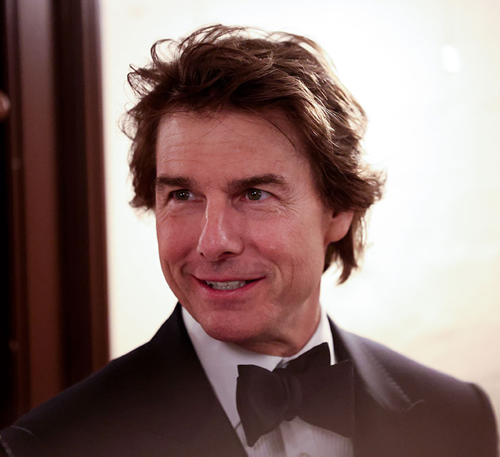 Tom Cruise in a tuxedo at an event, smiling and looking to the side with styled hair and soft lighting.