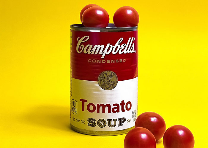 Campbell's tomato soup can with fresh tomatoes on top and beside it against a bright yellow background.