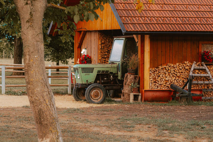 Green tractor parked beside a wooden shed with stacked firewood on a rural property in autumn.