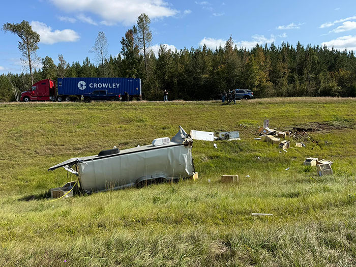 Wreckage of overturned trailer in grassy field with Crowley truck and people near treeline in background, heated debate online.