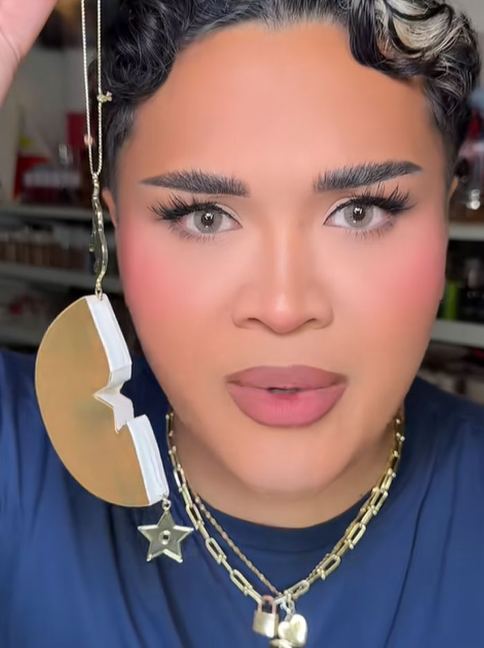 Person with styled hair holding a Dior advent calendar ornament, wearing layered gold necklaces and makeup.