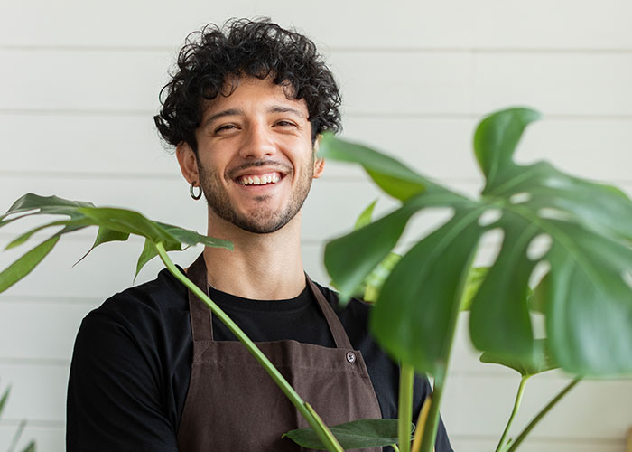 Smiling man wearing an apron standing behind large green leaves, capturing a moment of neighbor activities seen accidentally.
