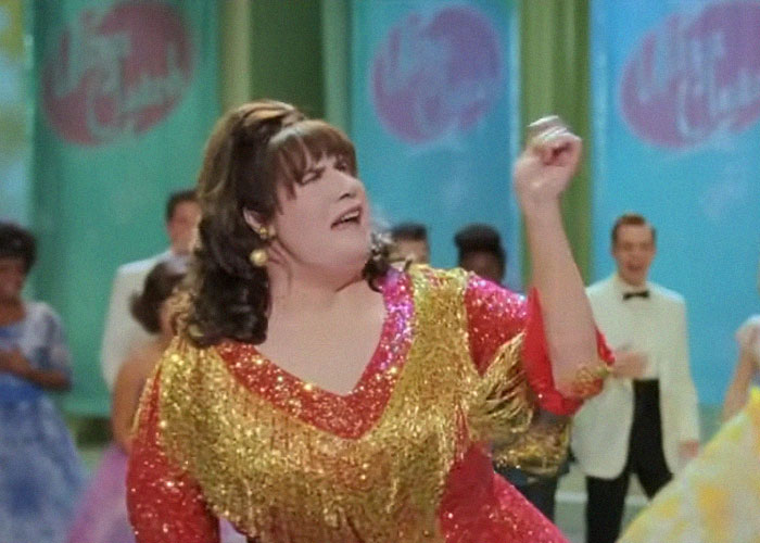 A person dancing energetically in a colorful sequined outfit during a lively community event with neighbors watching.