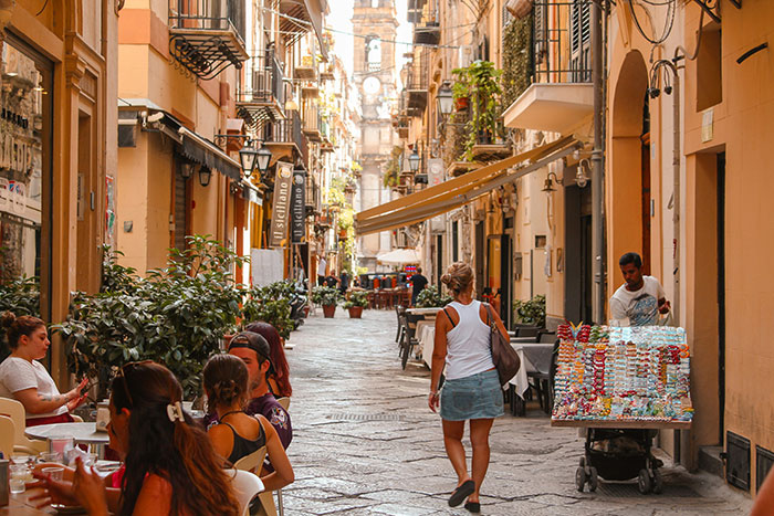 Solo female traveler walking down crowded narrow street in a European city with outdoor cafes and street vendor
