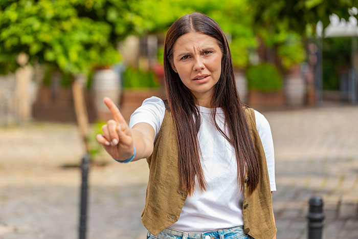 Young woman with long hair wearing a vest and white t-shirt making a stop gesture while walking outdoors.
