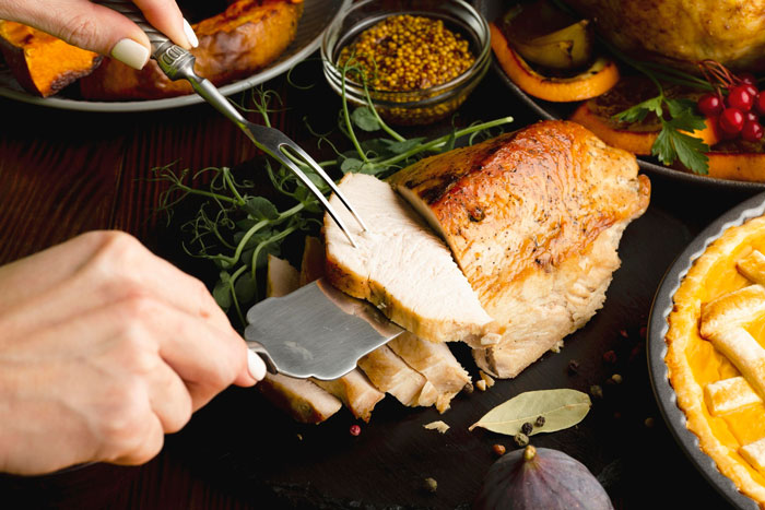 Person slicing a cooked roast on a platter surrounded by herbs, mustard, and autumnal side dishes. Person slicing a cooked roast on a platter surrounded by herbs, mustard, and autumnal side dishes.