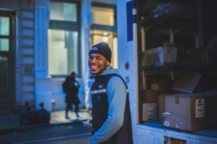 Young worker smiling outside delivery truck at dusk, sharing unprofessional things done while on the job.