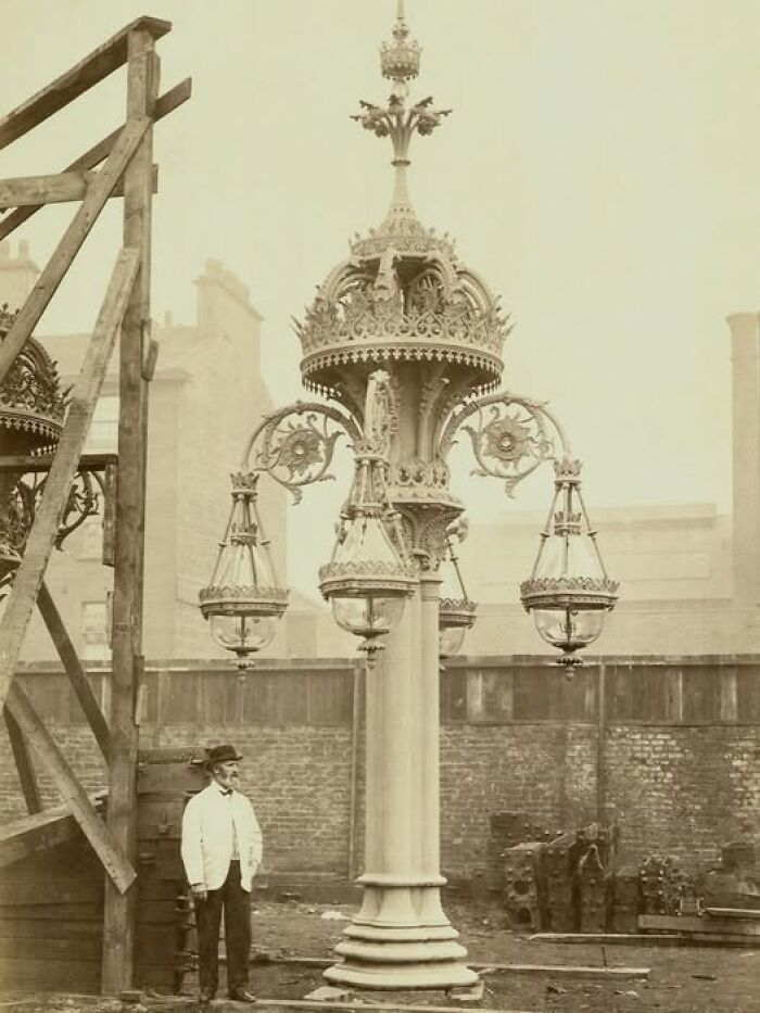 Man standing next to ornate Victorian-Edwardian street lamp post under construction in a sepia-toned outdoor setting