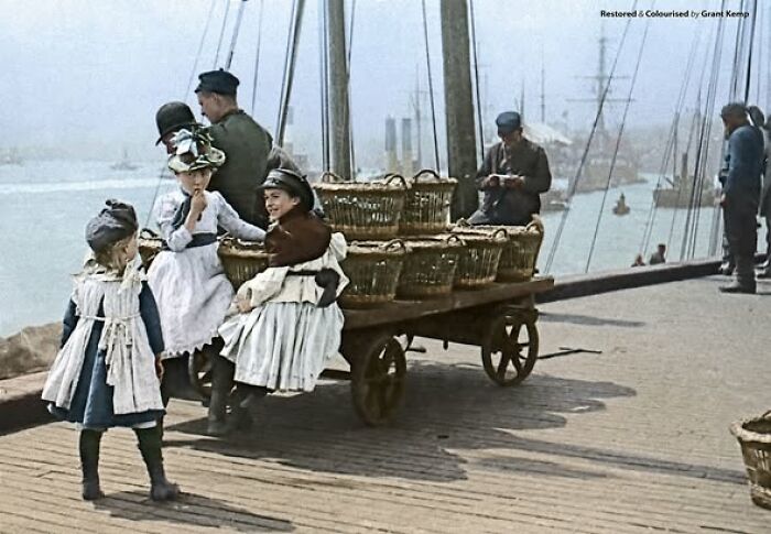 Children in Victorian-Edwardian pictures wearing period clothing by a dockside cart filled with baskets near boats.