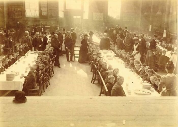 Victorian-Edwardian pictures of a large group of children and adults gathered for a meal in a spacious dining hall.