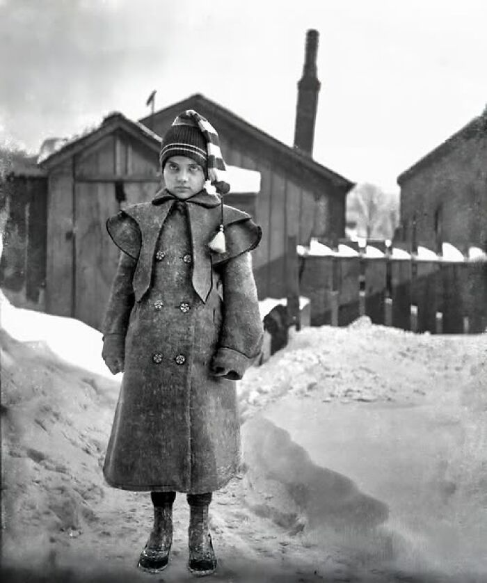 Young child wearing heavy winter coat and striped hat standing outside in snow, Victorian Edwardian pictures style.
