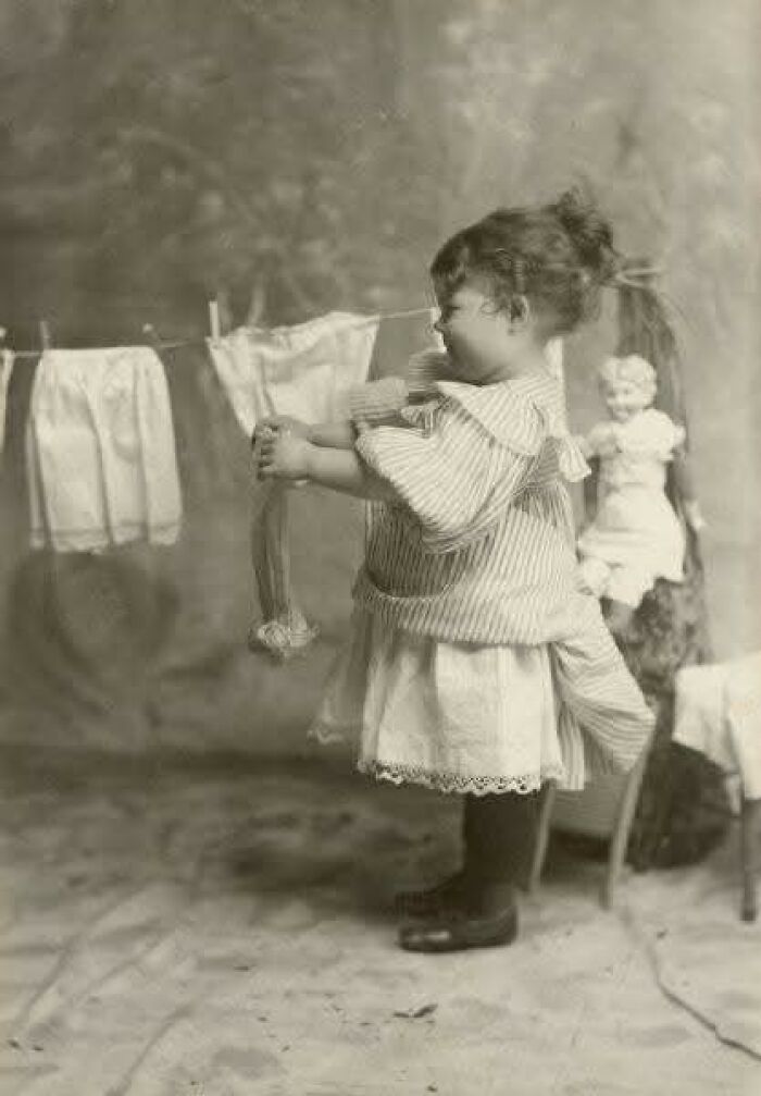 Young girl in Victorian-Edwardian pictures hanging doll clothes on a line indoors with vintage toys in background.