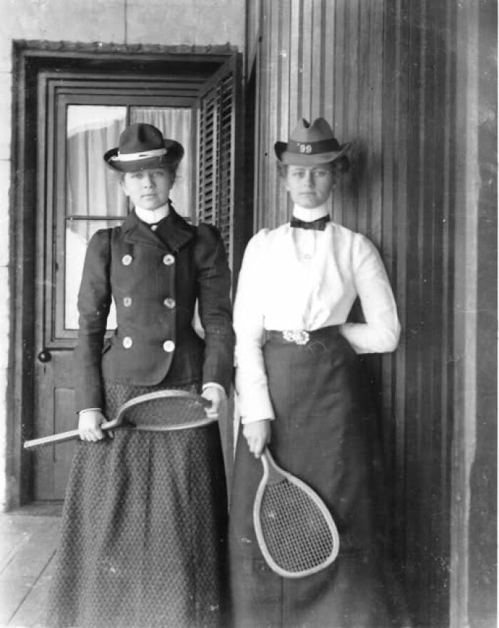 Two women in Victorian-Edwardian pictures wearing period clothing and hats while holding vintage tennis rackets.